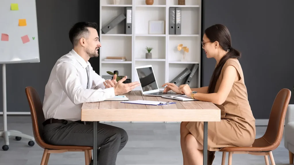 Two people sit across from each other at a wooden desk in a modern office. The man gestures while talking, and the woman types on a laptop. Documents and a plant are on the table; shelves and a whiteboard are in the background.
