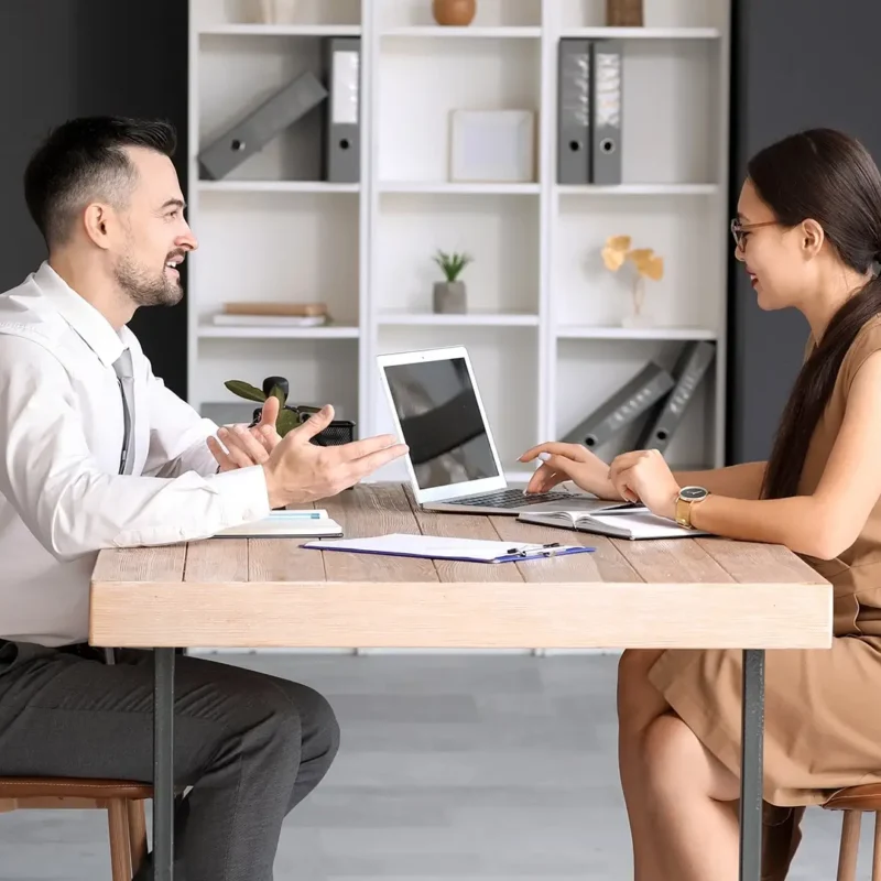 Two people sit across from each other at a wooden desk in a modern office. The man gestures while talking, and the woman types on a laptop. Documents and a plant are on the table; shelves and a whiteboard are in the background.