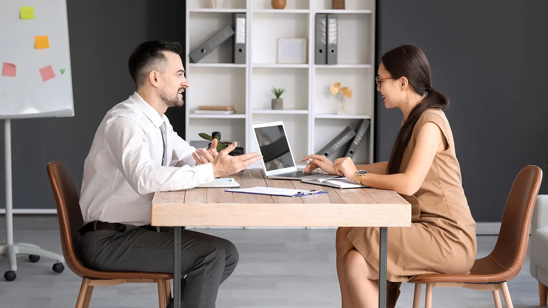 Two people sit across from each other at a wooden desk in a modern office. The man gestures while talking, and the woman types on a laptop. Documents and a plant are on the table; shelves and a whiteboard are in the background.