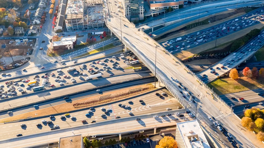 Aerial view of a busy highway interchange with multiple overpasses and heavy traffic; nearby construction sites, buildings, and autumn trees are visible.