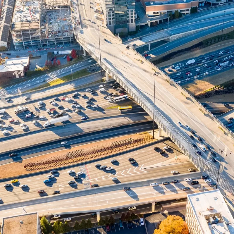 Aerial view of a busy highway interchange with multiple overpasses and heavy traffic; nearby construction sites, buildings, and autumn trees are visible.