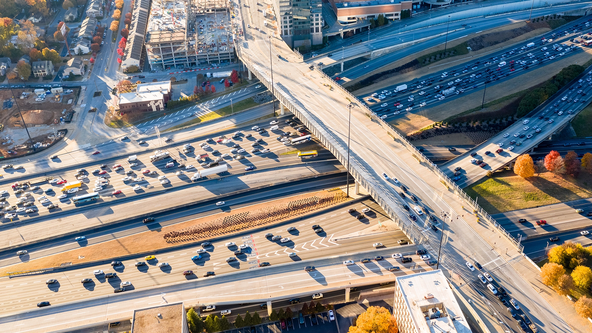 Aerial view of a busy highway interchange with multiple overpasses and heavy traffic; nearby construction sites, buildings, and autumn trees are visible.