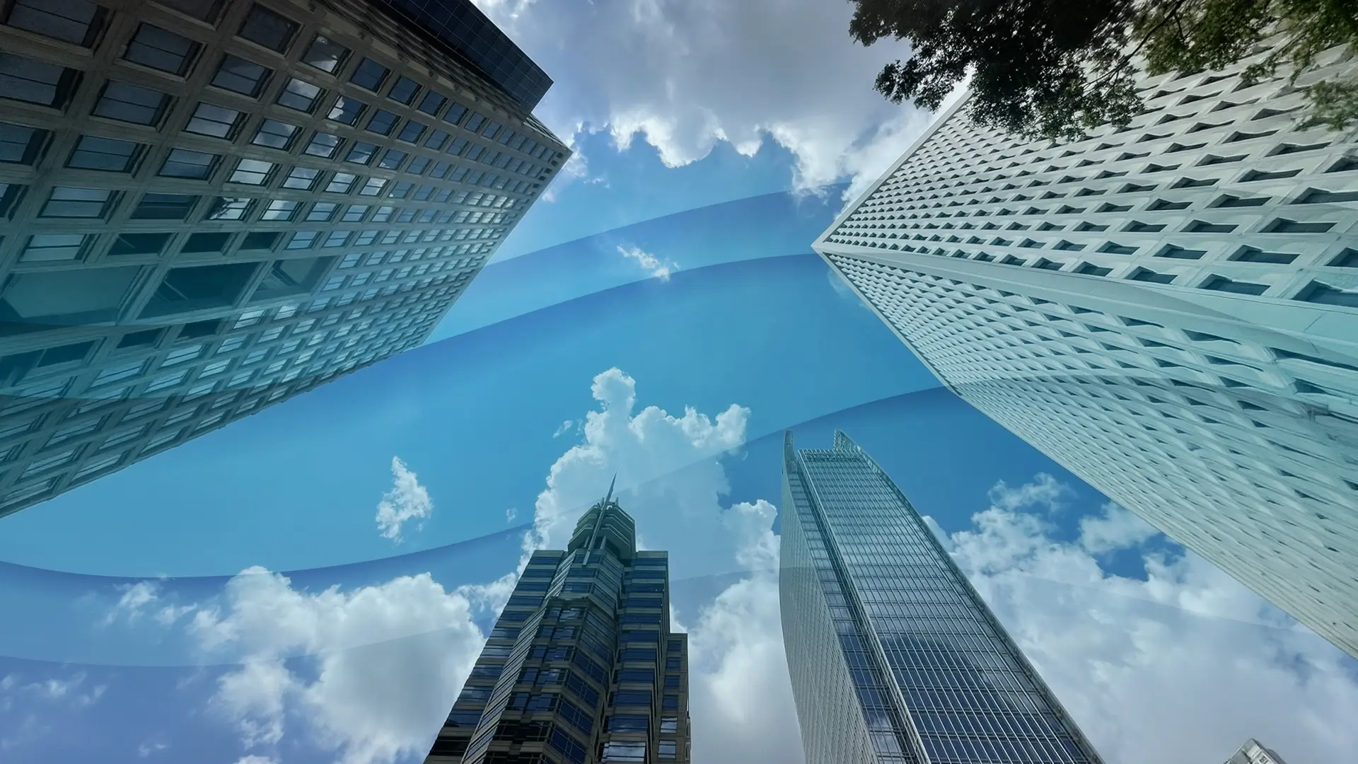 Four tall skyscrapers surround a bright blue sky with scattered clouds. The image has a surreal effect, with wavy blue bands cutting across the sky, distorting the view. Tree branches are visible in the top right corner.