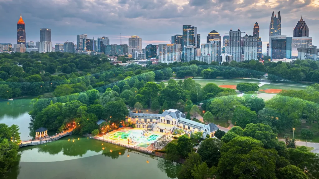 Aerial view of a city skyline with tall buildings, lush green trees, a small lake, a brightly lit pool area, and baseball fields at sunset under a cloudy sky.