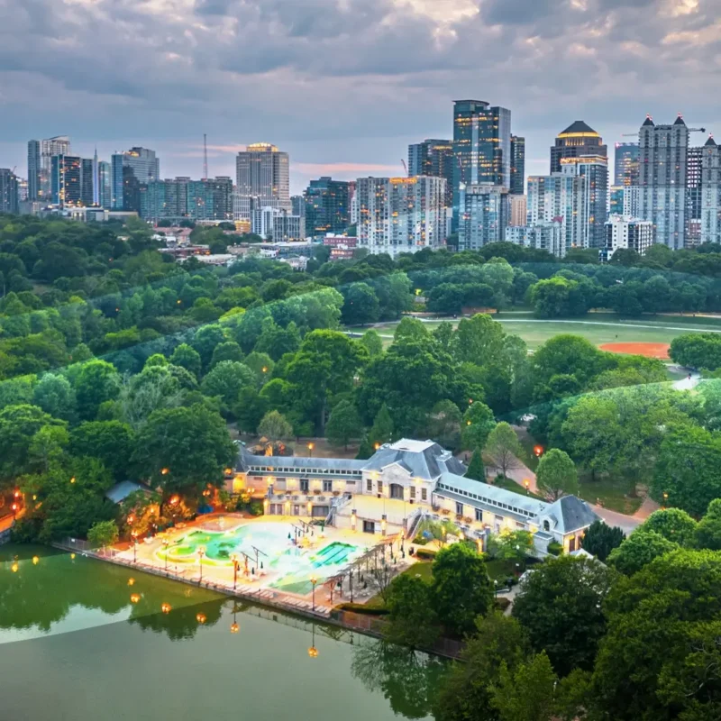 Aerial view of a city skyline with tall buildings, lush green trees, a small lake, a brightly lit pool area, and baseball fields at sunset under a cloudy sky.