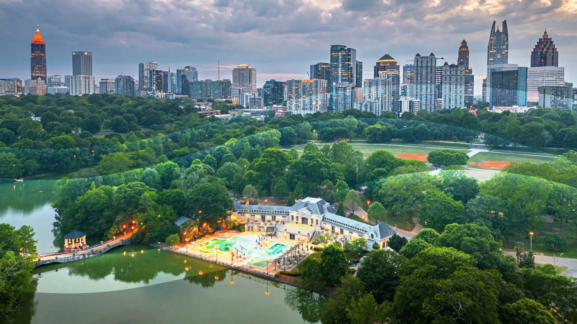 Aerial view of a city skyline with tall buildings, lush green trees, a small lake, a brightly lit pool area, and baseball fields at sunset under a cloudy sky.
