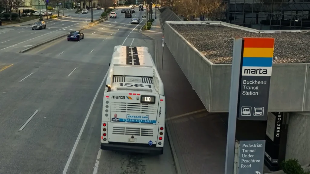 A MARTA bus numbered 1567 is stopped at the Buckhead Transit Station on a city street. Several cars are driving on the road, and a MARTA sign with station information is visible in the foreground.