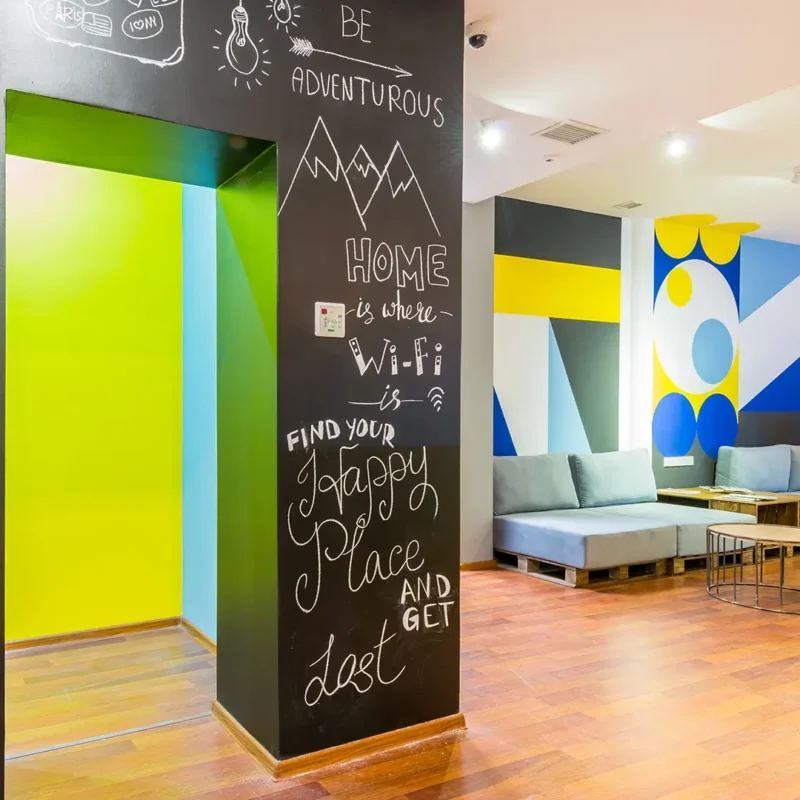 A modern lounge with colorful geometric wall art, blue sofas, wooden tables, and a black wall featuring motivational quotes written in white chalk. The space has wood flooring and bright lighting.