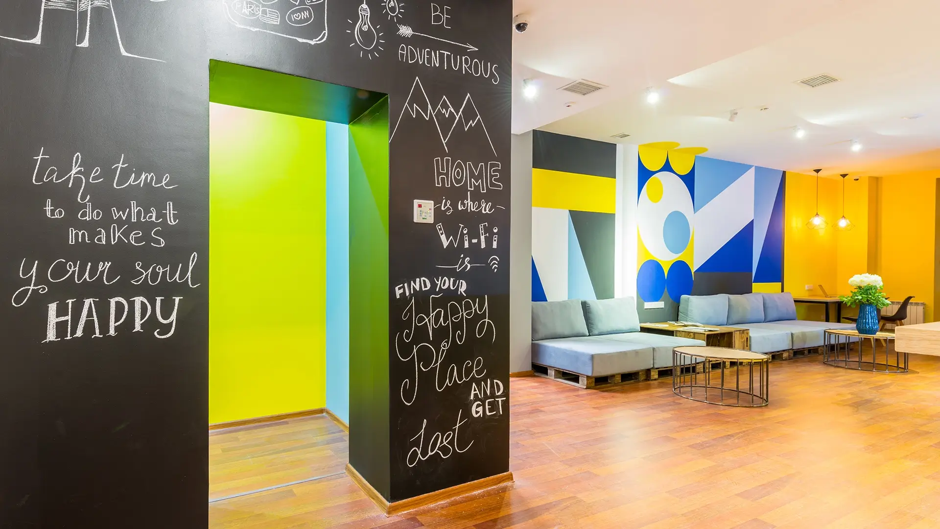 A modern lounge with colorful geometric wall art, blue sofas, wooden tables, and a black wall featuring motivational quotes written in white chalk. The space has wood flooring and bright lighting.