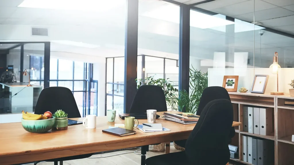 A modern office meeting room with a wooden table, four black chairs, notebooks, mugs, plants, and a bowl of fruit. Large windows let in natural light, and shelves with decor are visible in the background.
