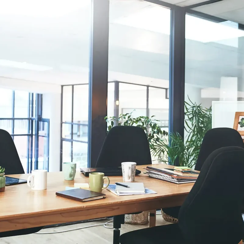 A modern office meeting room with a wooden table, four black chairs, notebooks, mugs, plants, and a bowl of fruit. Large windows let in natural light, and shelves with decor are visible in the background.
