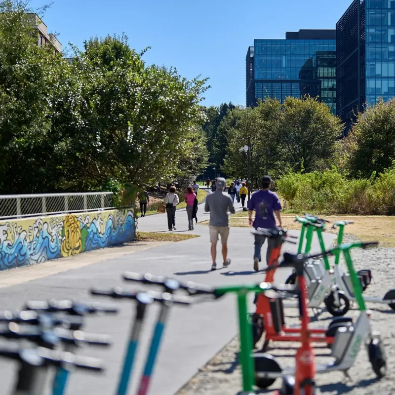 People walk and jog on a sunny urban path lined with trees and buildings, with several electric scooters parked in the foreground and colorful mural art on a nearby wall.