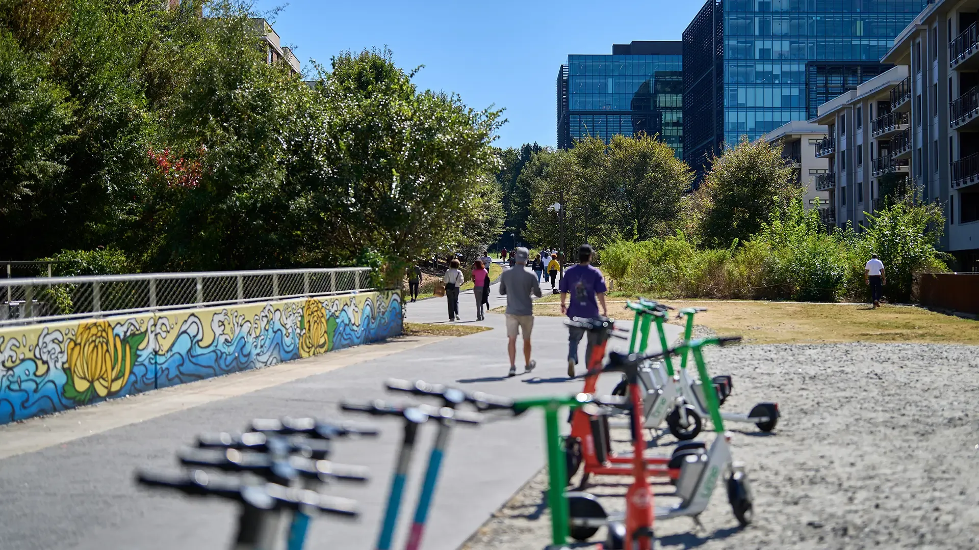 People walk and jog on a sunny urban path lined with trees and buildings, with several electric scooters parked in the foreground and colorful mural art on a nearby wall.