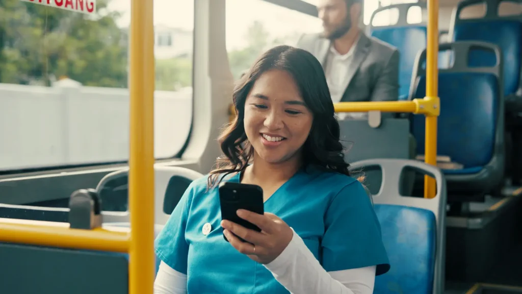 A woman in blue scrubs smiles while looking at her phone on a bus. Another person in business attire sits behind her. The bus has blue seats and yellow poles.