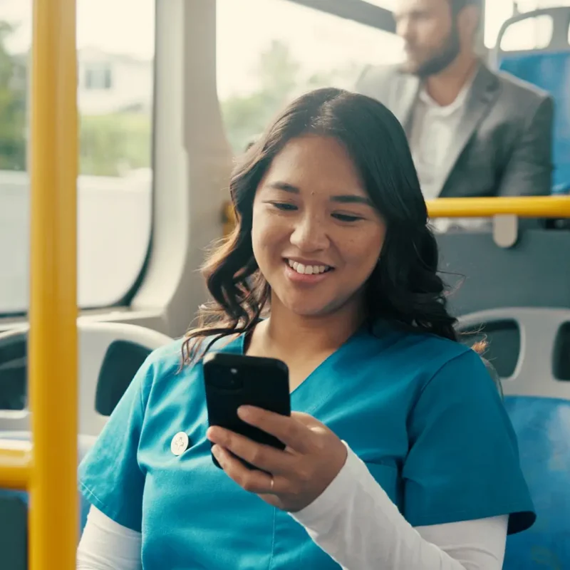 A woman in blue scrubs smiles while looking at her phone on a bus. Another person in business attire sits behind her. The bus has blue seats and yellow poles.