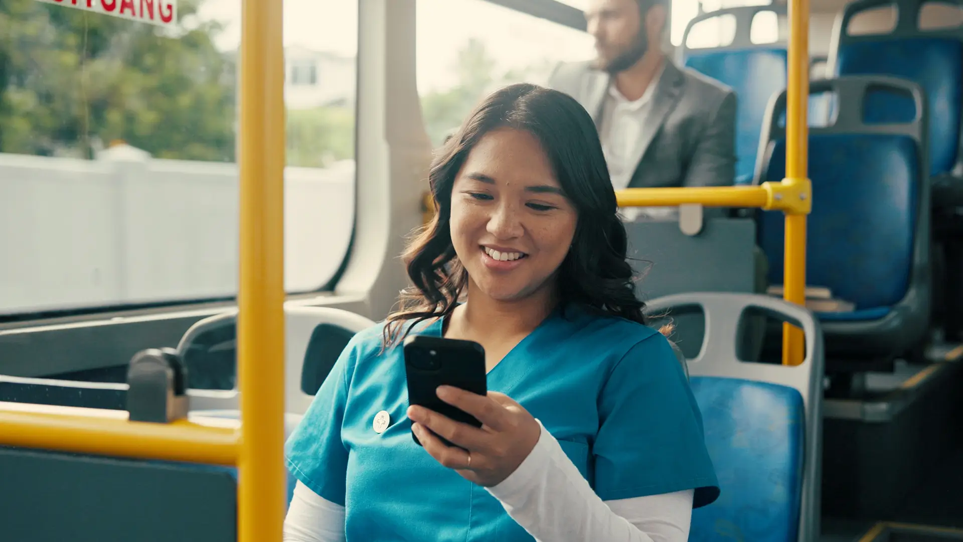 A woman in blue scrubs smiles while looking at her phone on a bus. Another person in business attire sits behind her. The bus has blue seats and yellow poles.
