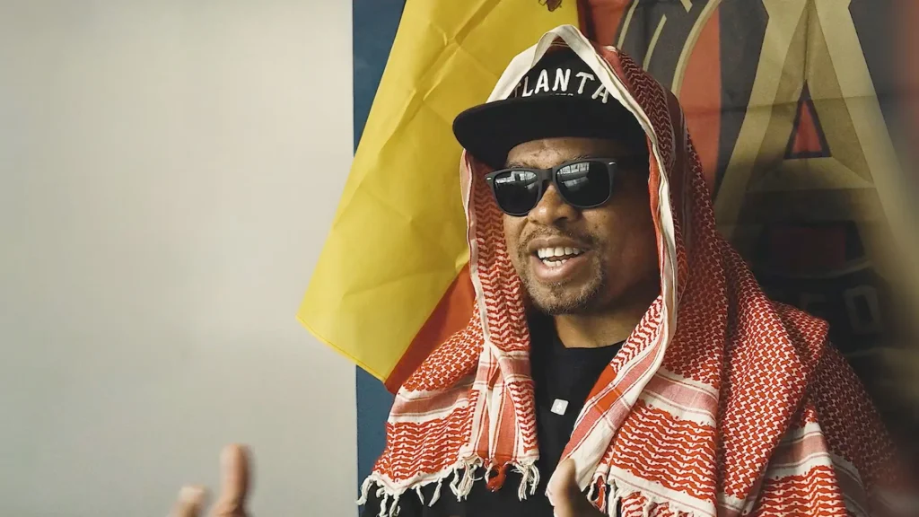 A man wearing sunglasses, an Atlanta baseball cap, and a red and white patterned keffiyeh.