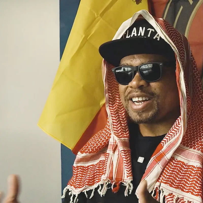 A man wearing sunglasses, an Atlanta baseball cap, and a red and white patterned keffiyeh.