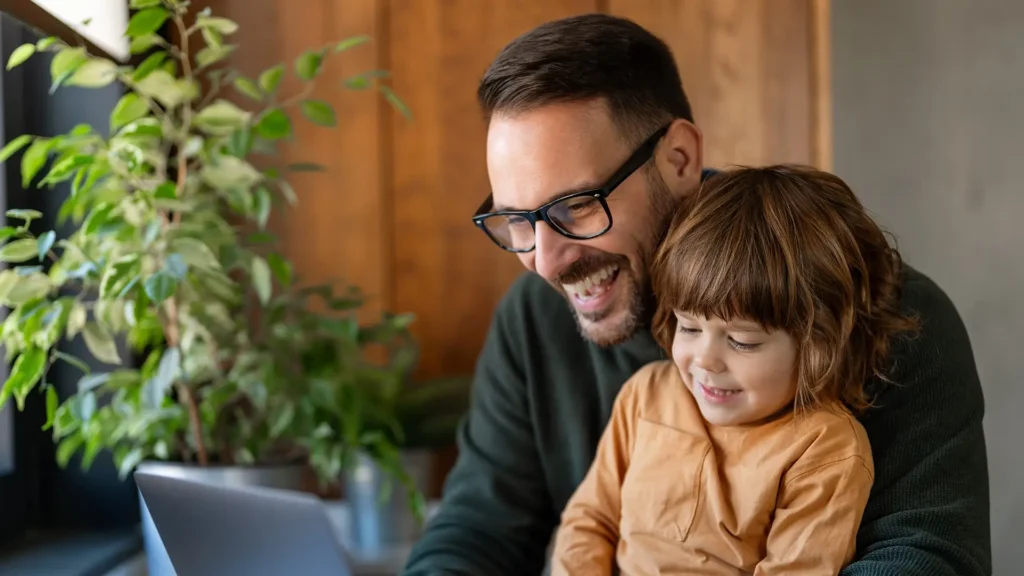 A smiling man with glasses sits with a young child on his lap while looking at a laptop screen.