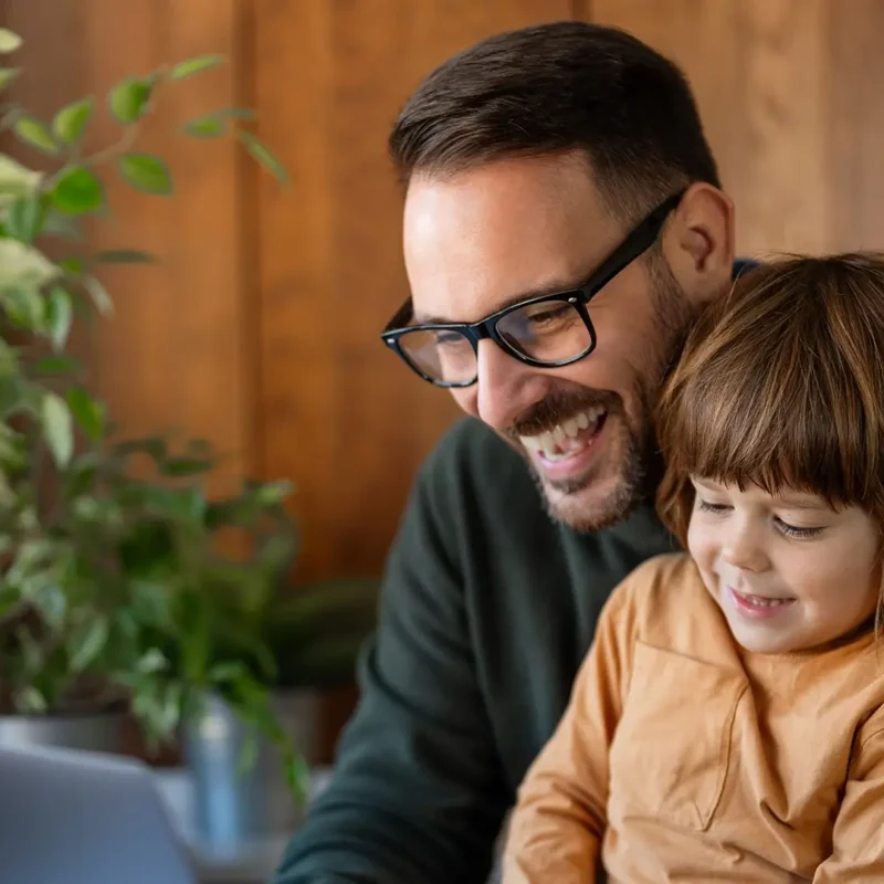 A smiling man with glasses sits with a young child on his lap while looking at a laptop screen.